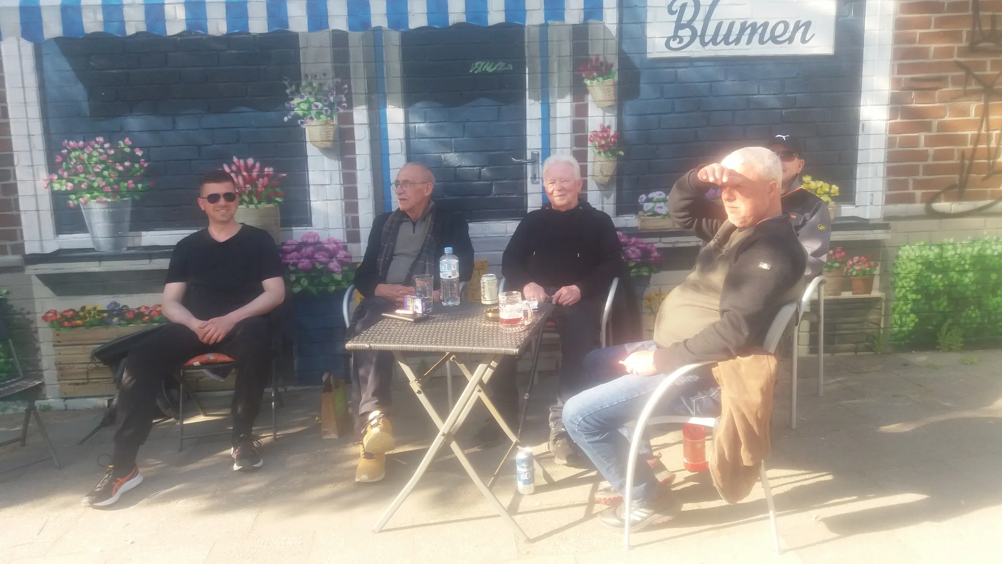 Vier Personen sitzen entspannt im Kaffeegarten von Bei Andy in Lübeck, an einem kleinen Tisch vor dem Kiosk mit blau-weiß gestreifter Markise und bunten Blumenkästen.