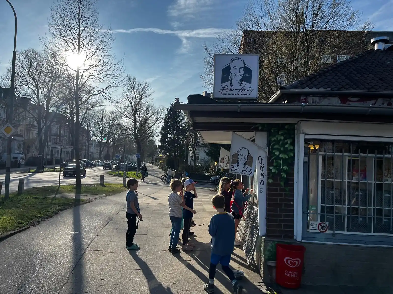 Kinder und Nachbarn stehen vor dem Kiosk Bei Andy im Lübecker Falkenviertel an einem sonnigen Frühlingstag, im Hintergrund Wohnstraße und blattlose Bäume