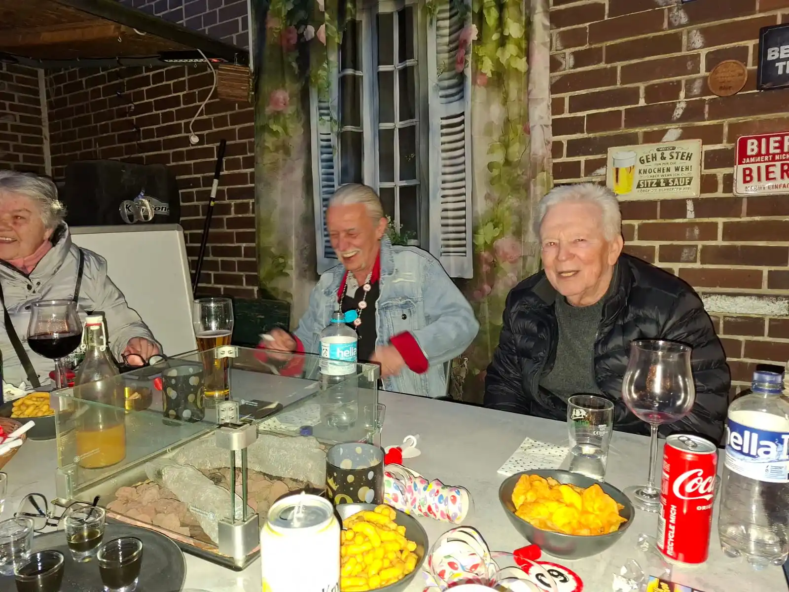 Drei ältere Herren sitzen lachend an einem Tisch im Kaffeegarten von Bei Andy in Lübeck, mit Grillgut, Getränken und Knabberzeug – typischer Grillabend-Moment im beliebten Nachbarschaftskiosk.