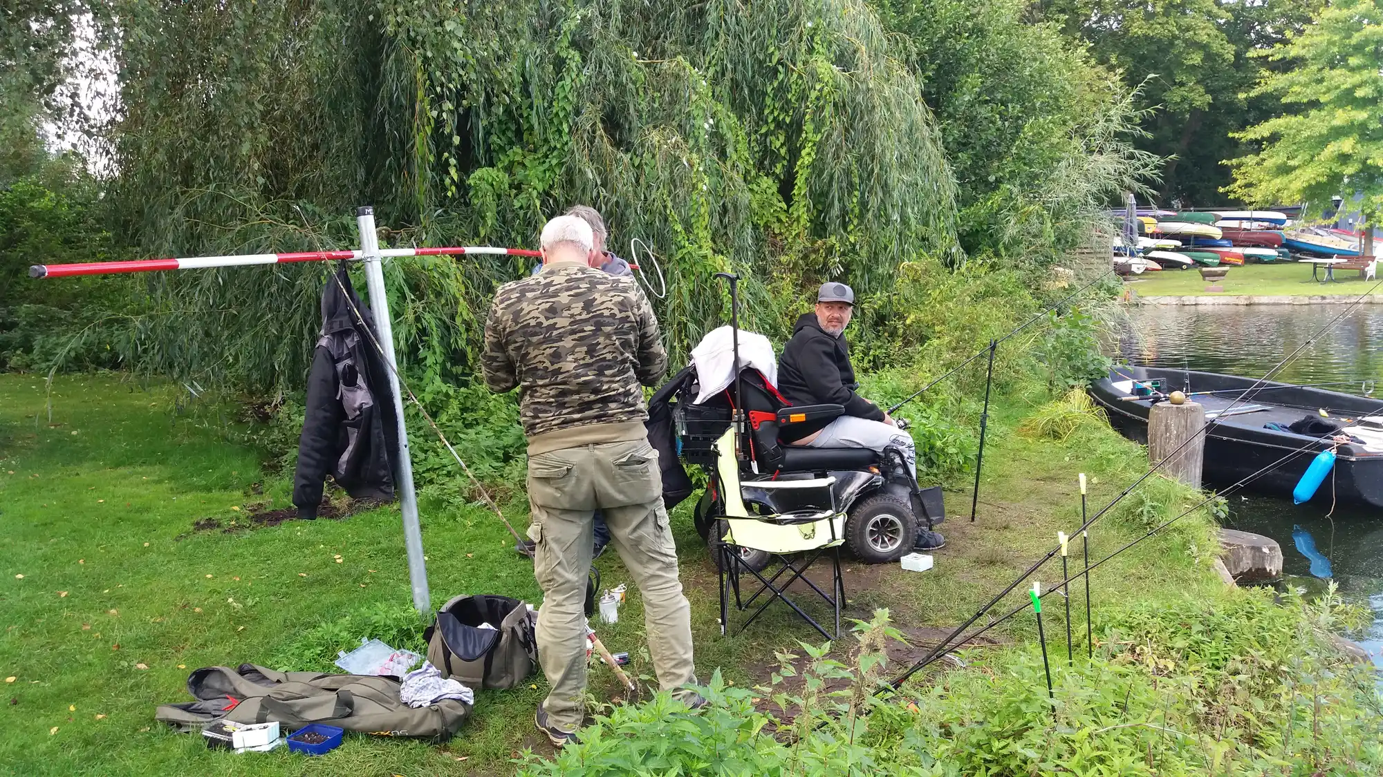 Besucher beim Grillabend im Kaffeegarten Bei Andy an der Wakenitz in Lübeck: Mann mit Grill unter Segeldach, Gäste sitzen entspannt auf der Wiese, Boote im Hintergrund.