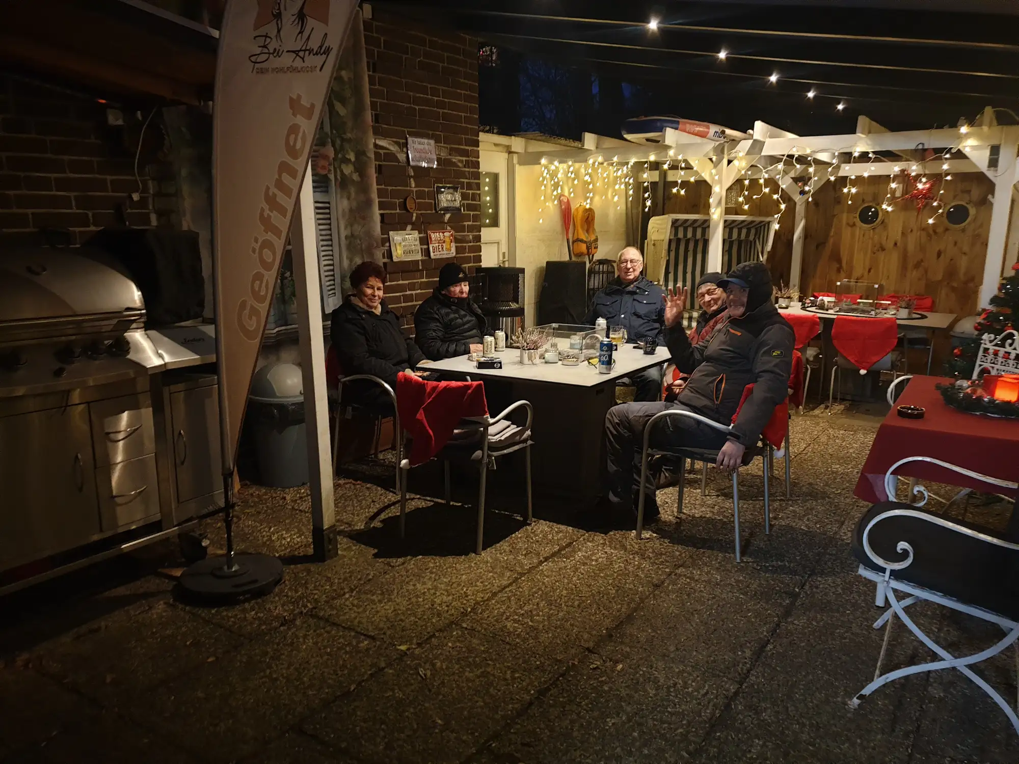 Fünf Personen sitzen gemütlich um einen Tisch im Kaffeegarten Bei Andy in Lübeck, im Hintergrund weiße Pergola mit Lichterketten und Grillgeräte, warme Abendatmosphäre.