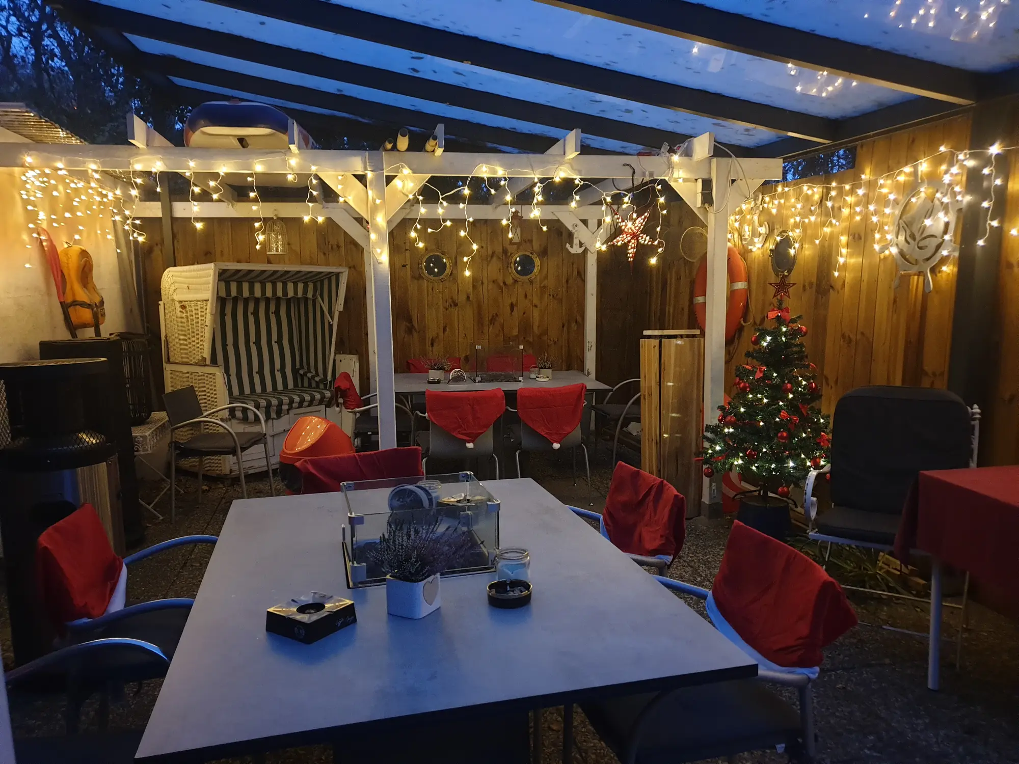 Festlich geschmückter Kaffeegarten Bei Andy in Lübeck mit warmweißen Lichterketten, roten Stuhlauflagen, Weihnachtsbaum und gemütlichen Holztischen unter weißem Pergola-Dach