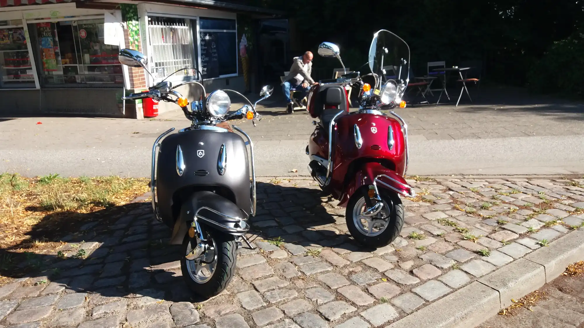 Zwei bunte Vespa-Roller, einer schwarz und einer rot, parken im sonnigen Kaffeegarten von Bei Andy am Wakenitz in Lübeck