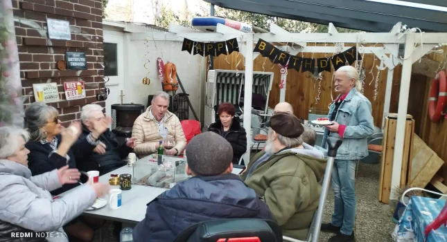 Gruppe von Stammkunden sitzt beim Geburtstagsfest im Kaffeegarten Bei Andy in Lübeck zusammen, im Hintergrund ein Happy-Birthday-Banner und rustikale Holzwände