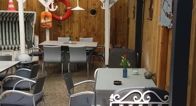 Gemütlicher Kaffeegarten Bei Andy in Lübeck mit schwarzem Pergola-Dach, Holzwänden, Steintischen und nostalgischen Dekoelementen unter grünen Bäumen